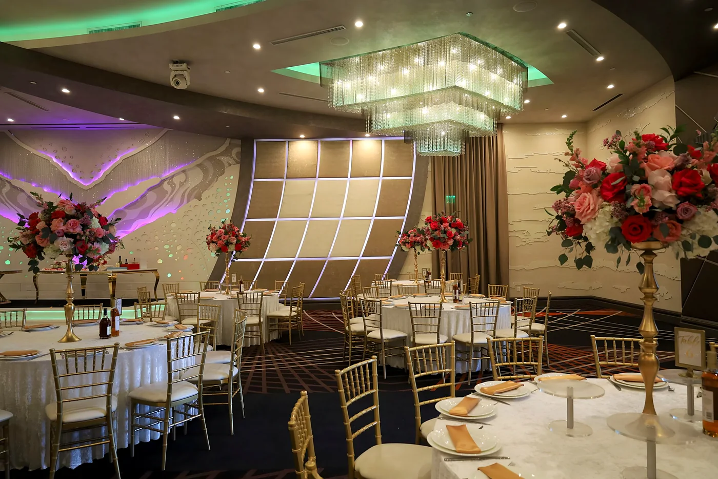 Ballroom with gold chairs, a crystal chandelier and tall red and blush rose centerpieces on gold stands