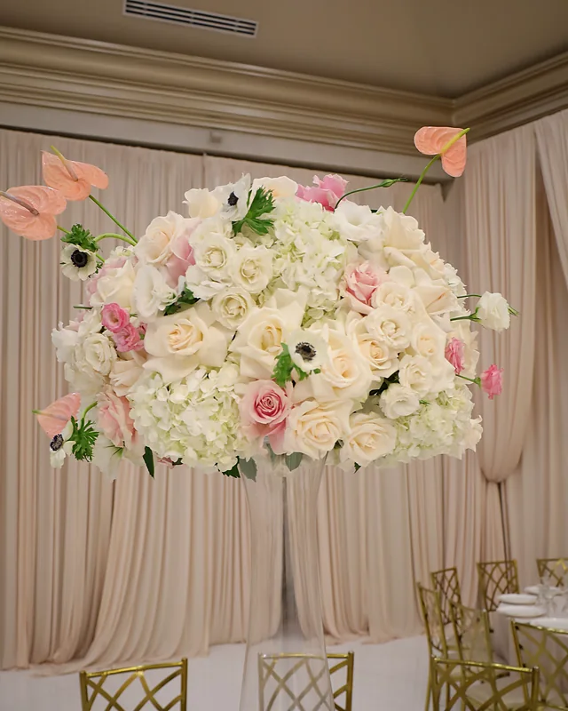 Domed centerpiece of cream roses, white hydrangea, anemones and coral anthurium against ivory drapes
