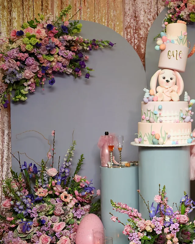 Tiered bunny first birthday cake surrounded by lavender, pink and purple spring flowers and candles