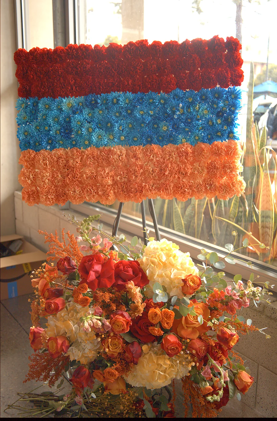 Floral Armenian flag made of orange, blue and red carnations and daisies, beneath a large arrangement of roses and hydrangea